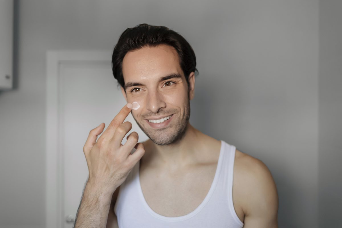 A Handsome Young Man Applying Moisturizing Cream on His Face While Standing in Front of the Mirror. Skin Care and Beauty Concept.