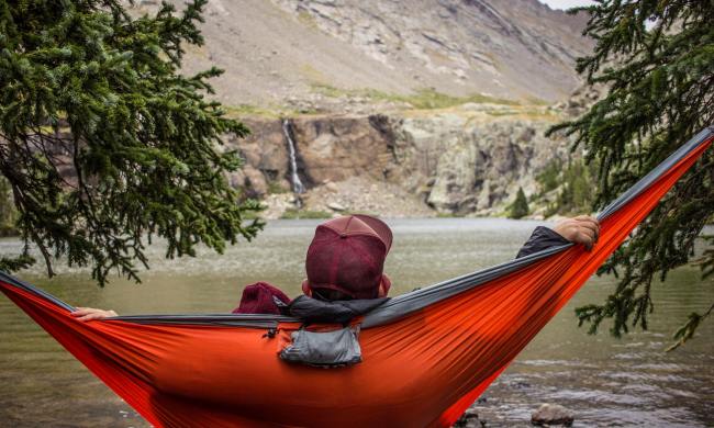 A man relaxing on a red camping hammock by a body of water.