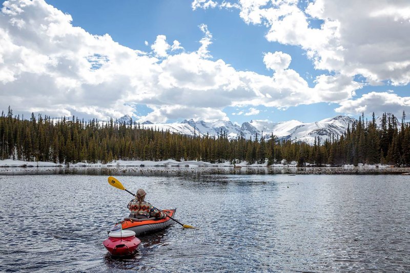 Kayaker paddling an inflatable kayak with a floating cooler in tow and snowy mountains in the background.
