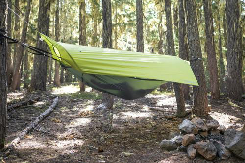 A green Klymit hammock tent strung between two tress in a forest.