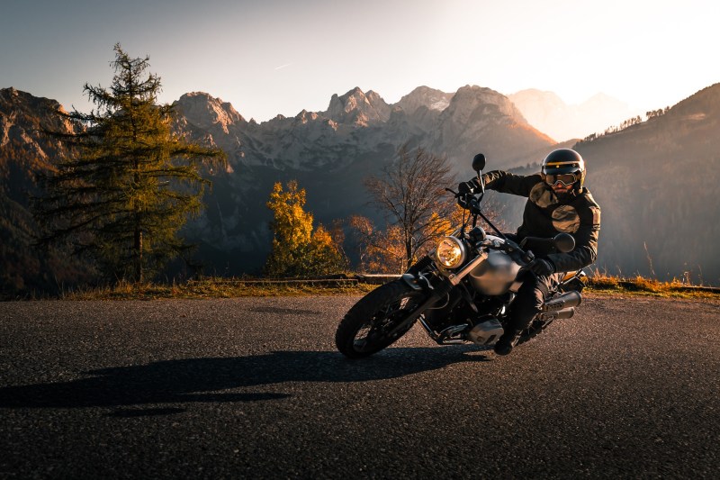 Motorcycle rider leaning into a corner on a mountain road with the sun and mountains in the back.