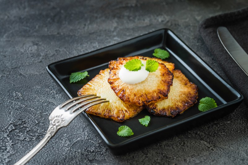 Fried pineapple slices with creme fraiche, mint in black square plate on dark background
