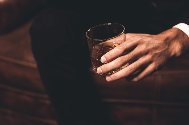 A close up of a man's hand holding a highball glass with about two fingers of whiskey in it. 