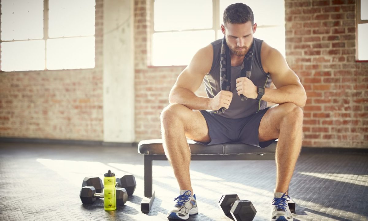 A tired man with towel sitting on a bench in a gym.