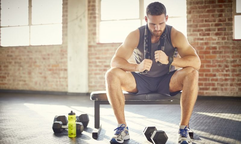 A tired man with towel sitting on a bench in a gym.