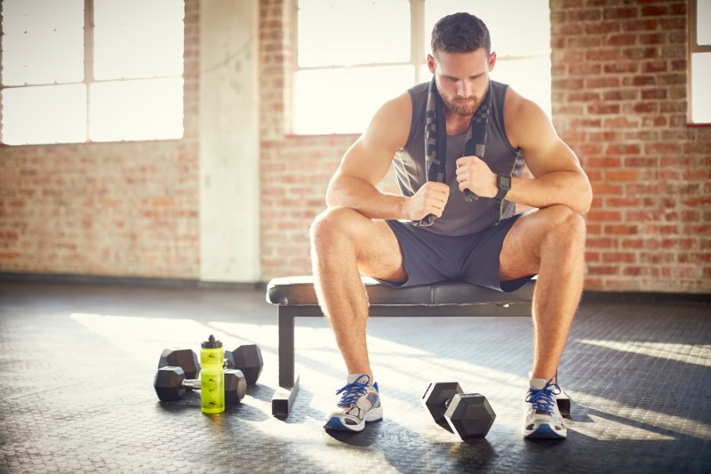 A tired man with towel sitting on a bench in a gym.
