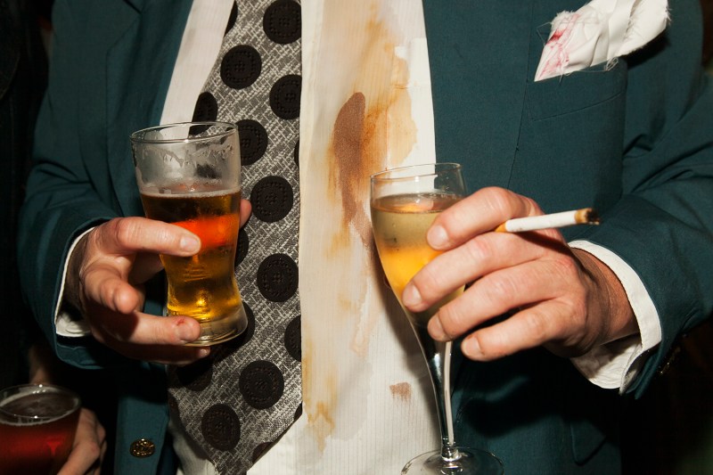 Midsection of a man holding drinks and cigarette at party with a beer stain on his shirt.