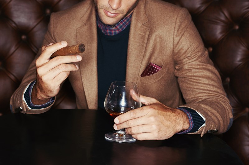 A stylish man drinking whiskey and smoking a cigar.