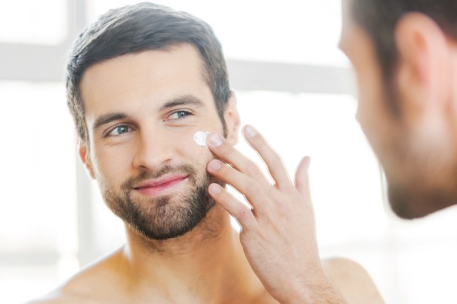 Handsome young shirtless man applying cream at his face and looking at himself with smile while standing in front of the mirror.