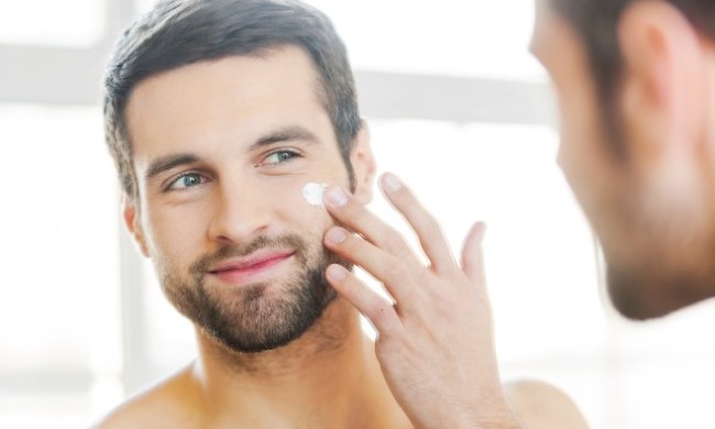 Handsome young shirtless man applying cream at his face and looking at himself with smile while standing in front of the mirror.