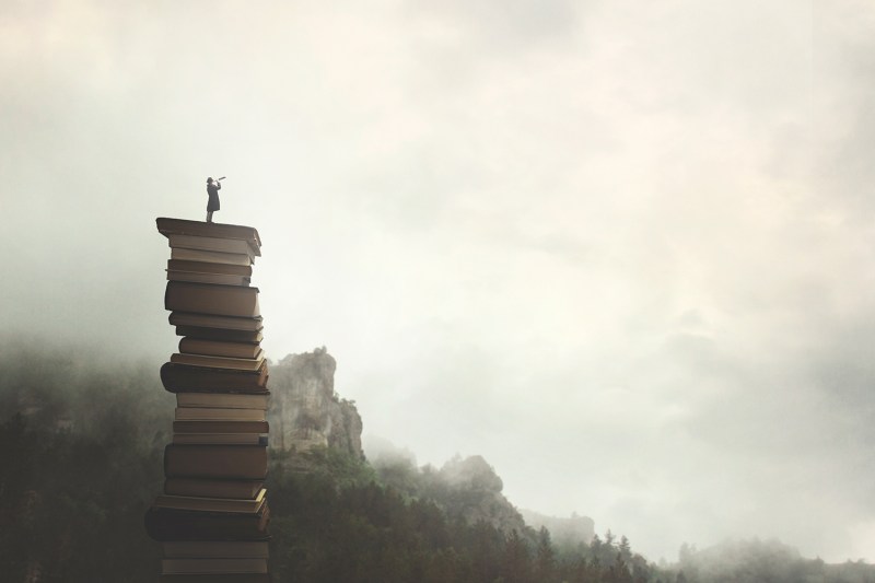 surreal man with telescope looks at infinity from the top of a stack of books in the outdoors.