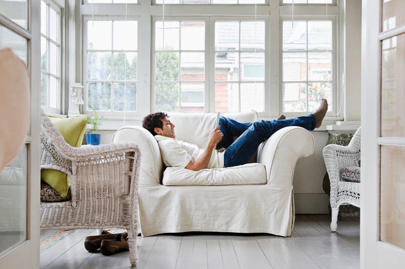 Man laying down on couch reading book next to window.