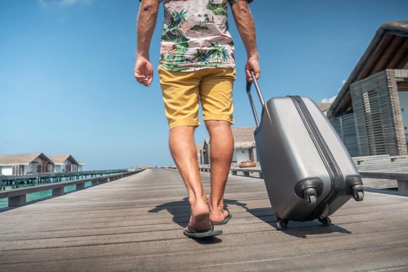 A man wearing a Hawaiian shirt and yellow shorts is seen pulling his luggage to his hotel room in a tropical destination. 