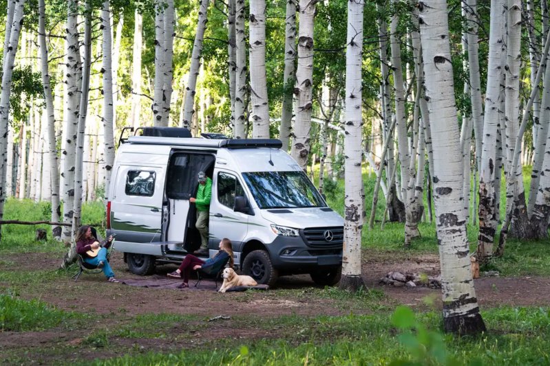 Three campers and a dog relaxing at a wilderness campsite with a Winnebago Revel campervan.