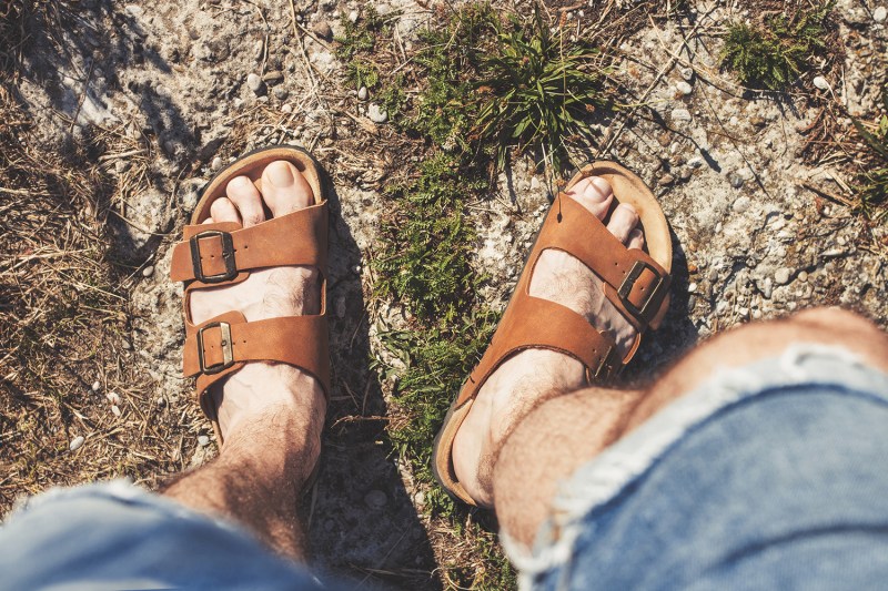 Downward view of a man's feet wearing brown sandals standing on grass.