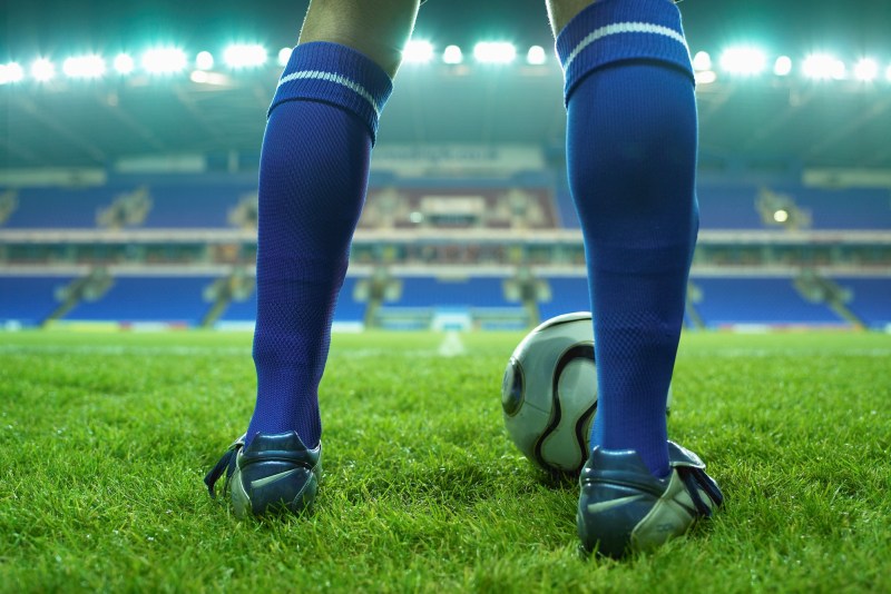 A close-up shot of men's legs standing on a grassy field, wearing blue knee-high socks with a ball at the front of his right foot.