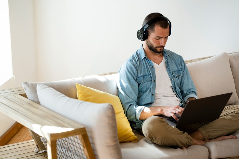 Young man sitting on couch at home with headphones and laptop.