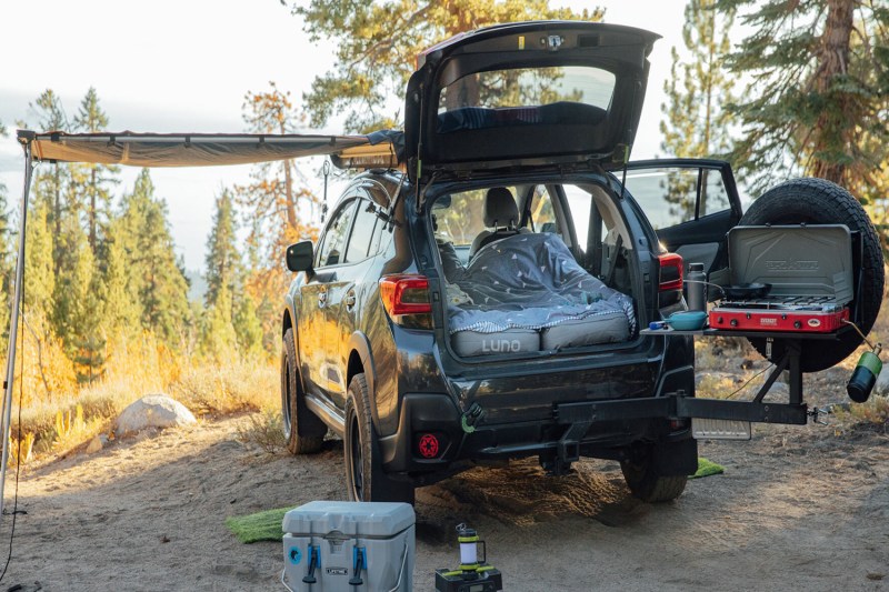 A vehicle with a blanket and Luno air mattress, camping stove, camping lantern, and cooler in a campsite.