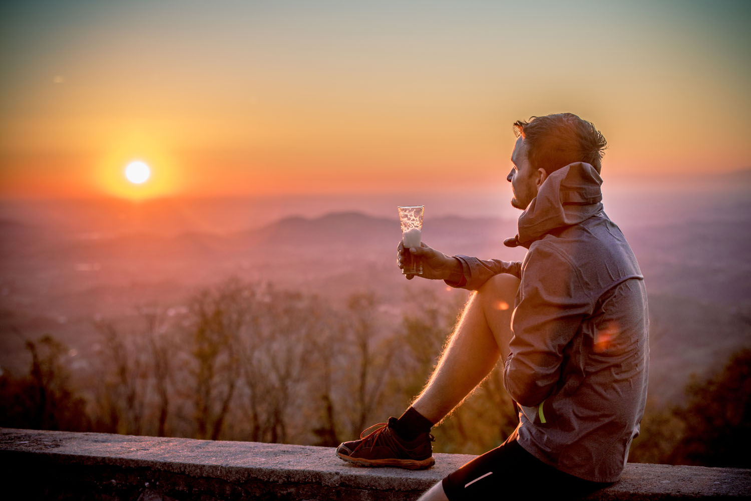 A man sitting on a wall and holding a glass of beer by the sunset.