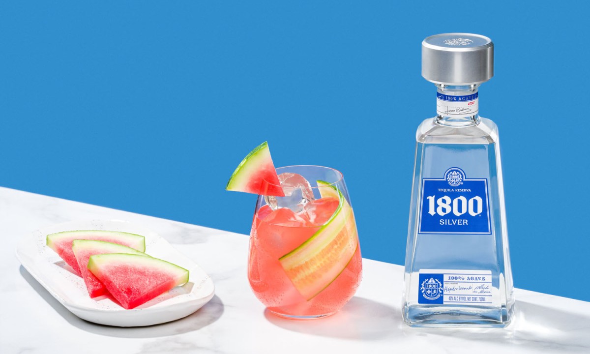 A glass of Summer Sangria in between a plate of watermelon slices and a bottle of 1800 Silver Tequila.