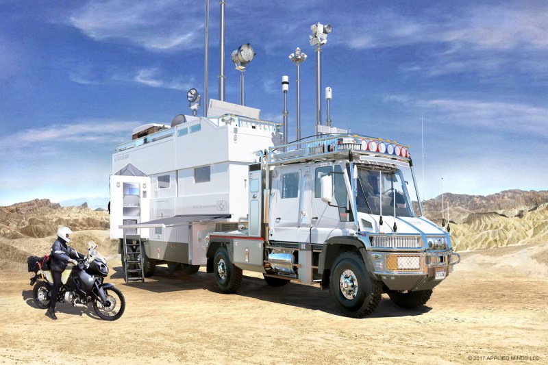 Rider on a motorcycle beside the Applied Minds KiraVan Expedition Systems overlanding vehicle.