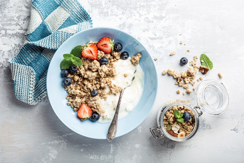 A blue towel, a bowl of yogurt with granola, blueberries, strawberries, and a mint leaf, and a jar of granola on a table.