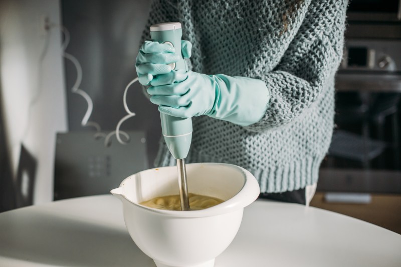 hands with protective gloves using a hand blender on a white bowl.