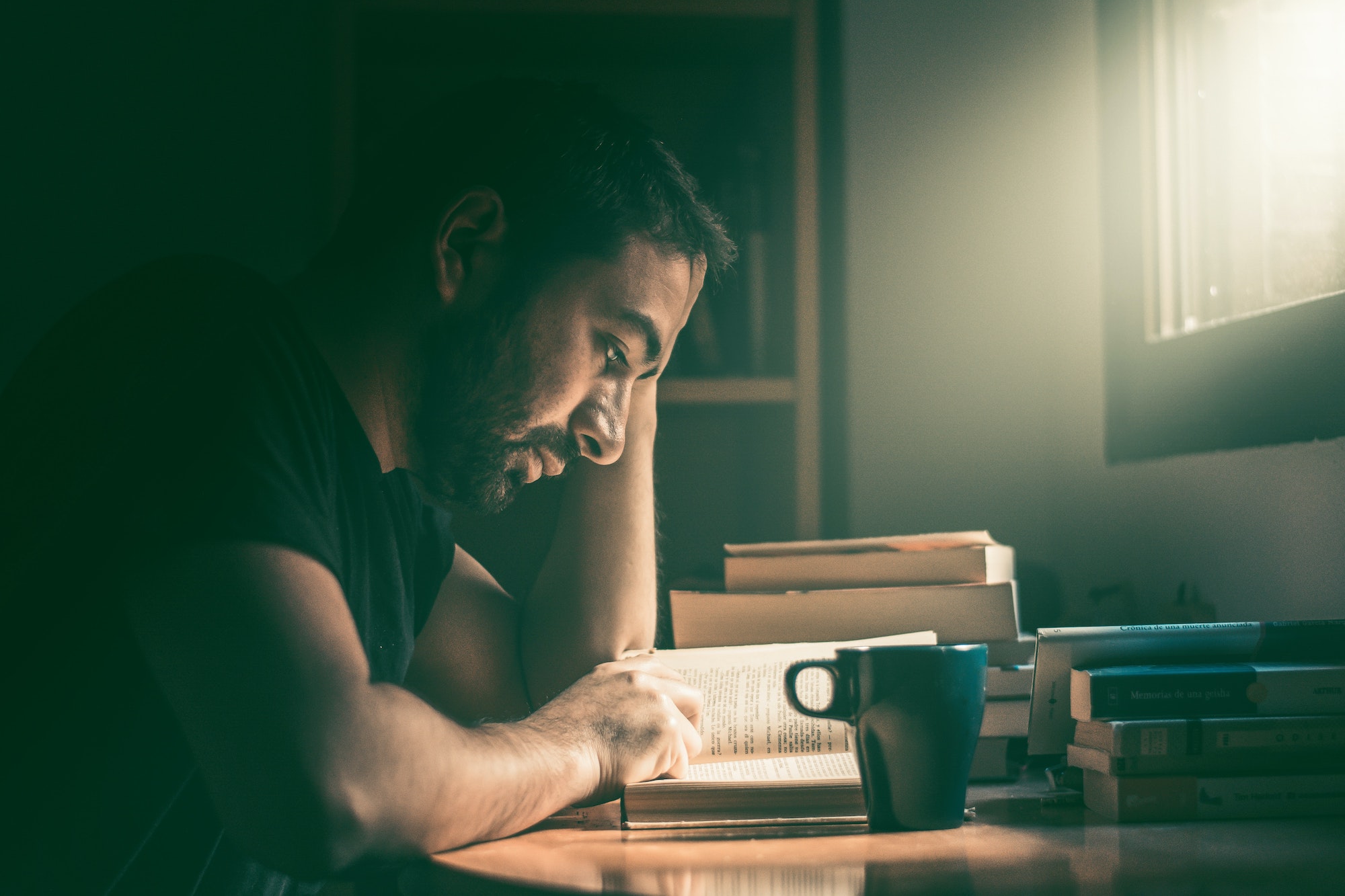 Young man reading a novel with a cup of coffee next to stack of books.