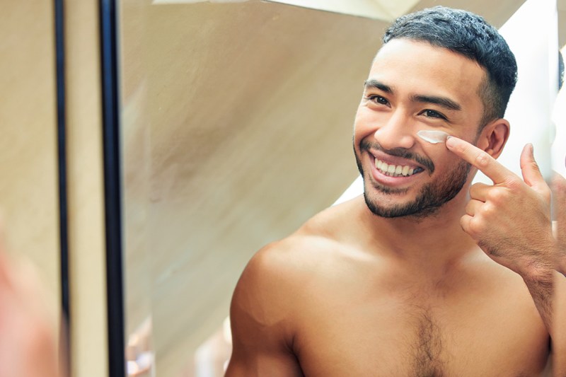 Shot of a young man looking in the mirror while applying moisturizer to his face.