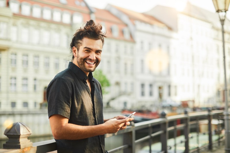 A smiling man in front of a landmark is seen holding a phone and wearing a black short-sleeve shirt.