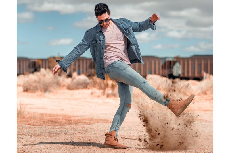 man in denim and desert boots kicking up sand.