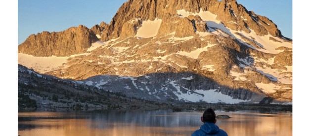 Man facing an alpine lake as the sunrises on a mountain peak.