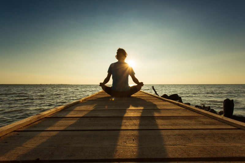 Young man sitting and meditating on a pontoon by a lake at the sunrise.