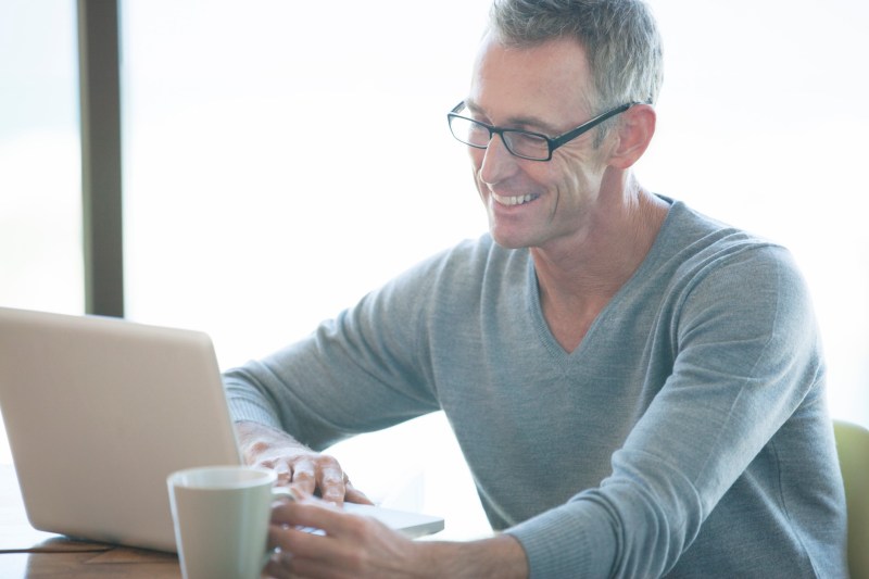 Man wearing a v-neck sweater while using a laptop and having a cup of coffee.