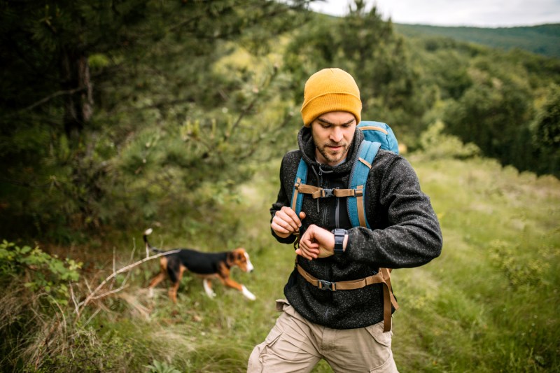 A male hiker checking the time on his watch as a dog walks behind him.