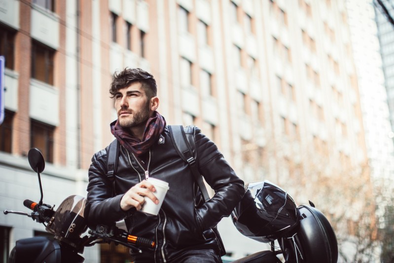 Handsome modern man enjoys a lovely day out in the city. He is holding a cup of coffee while sitting on a motorcycle.