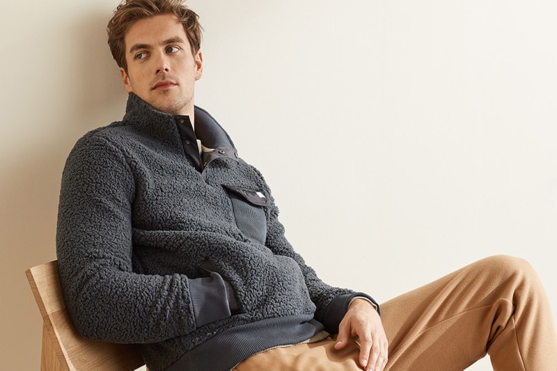 A male model in sweatshirt and pants sitting on a chair.