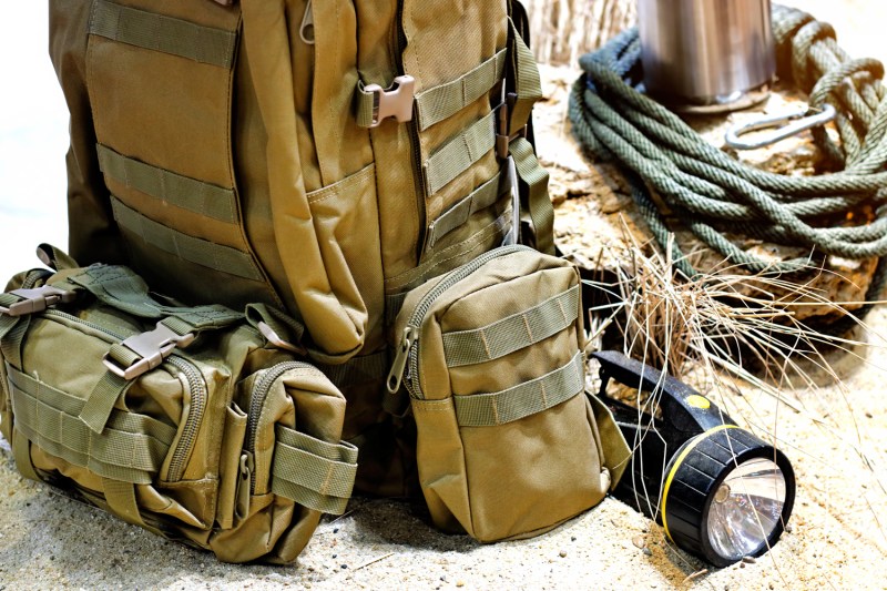 A tactical backpack beside a flashlight and rope on sand.