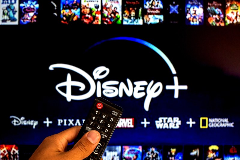 Close-up of a hand holding a remote with a TV displaying the Disney Plus app in the background. 