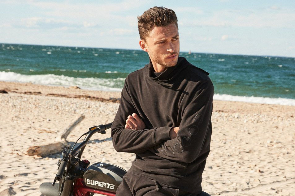A man in a black long-sleeved shirt sits on a motorcycle on the beach with the ocean behind him.