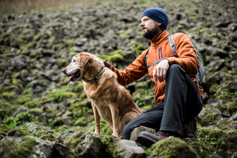 A crouching male hiker and his dog on mossy rocks.