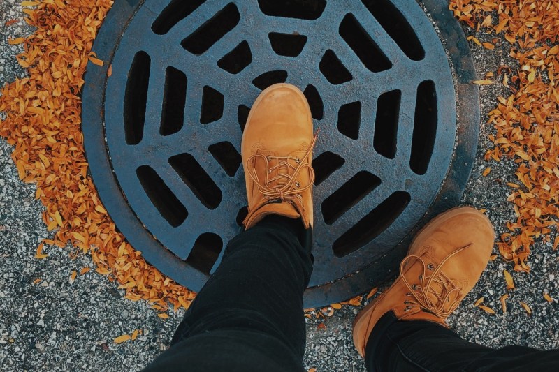 Person wearing Timberland boots while standing on a street drain surrounded by leaves