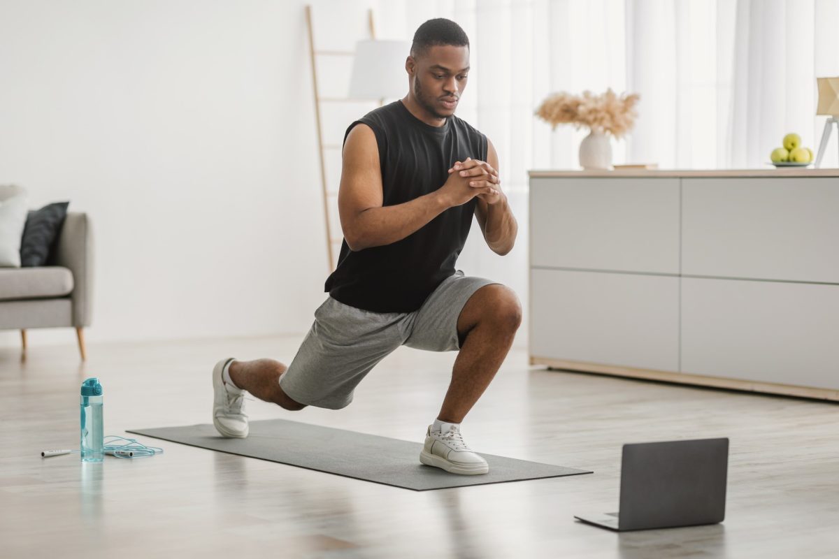Black man doing alternating forward lunges in front of a laptop at home.