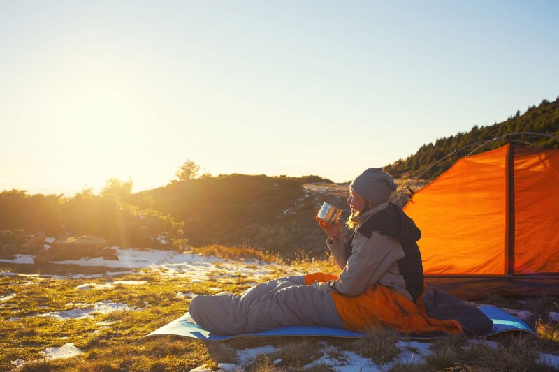 A woman camping outdoors sitting in her sleeping bag and sipping a drink. 