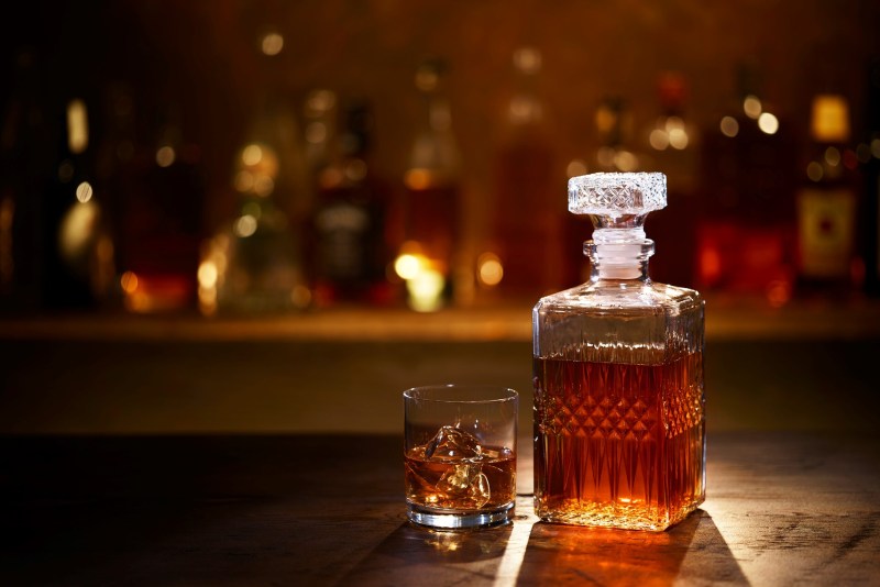 A Bourbon Whiskey Decanter and Bourbon on the Rocks in a Tumbler sitting on a bar with bottles in the background.