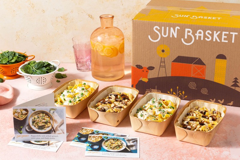 Recipe cards and four sets of healthy meals in front of a Sun Basket box, with flavored water and veggie leaves beside them.