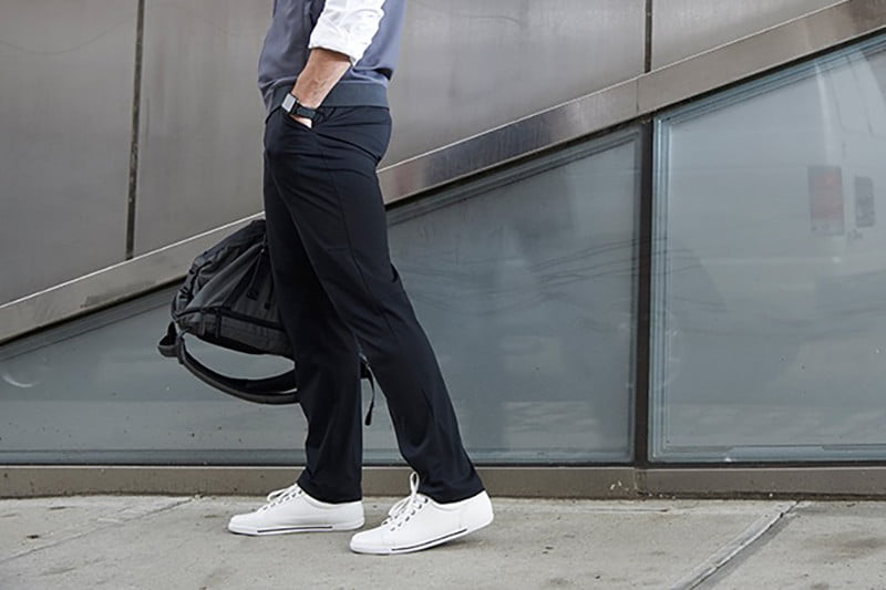 lower half of a man holding a duffle bag and wearing black pants with white sneakers.