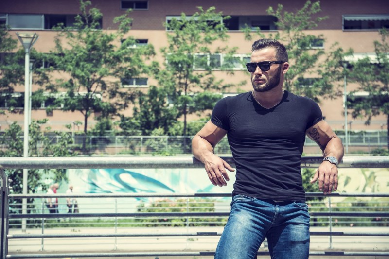 A muscular man in a black t-shirt and blue jeans leaning against a fence.