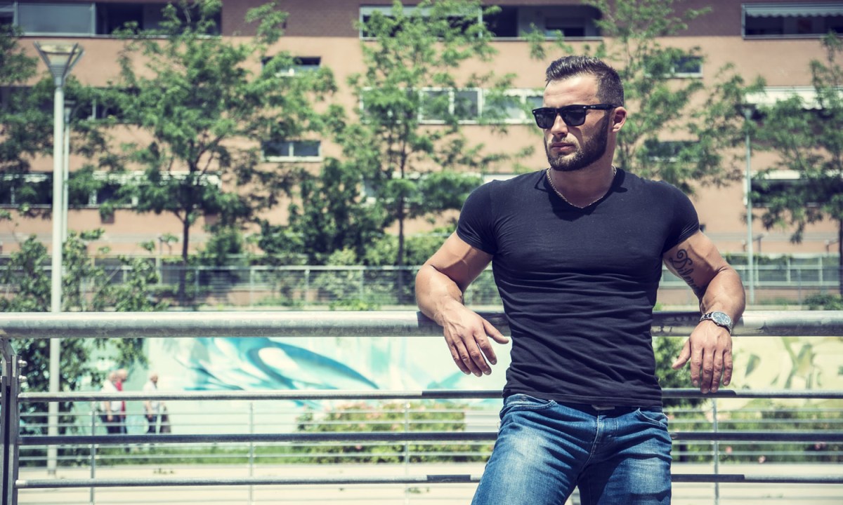 A muscular man in a black T-shirt and blue jeans leaning against a fence.