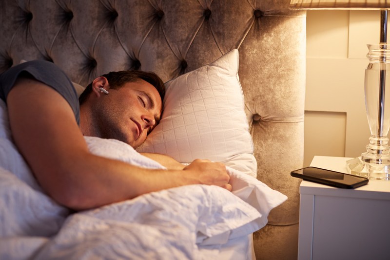 Man Sleeping In Bed Wearing Wireless Earphones Connected To Mobile Phone On Bedside Table.